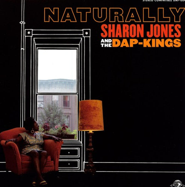 Cover art for Sharon Jones And The Dap-Kings album 'Naturally' featuring a photograph of Jones sitting in a chair with a lamp and a view of a building through the window.