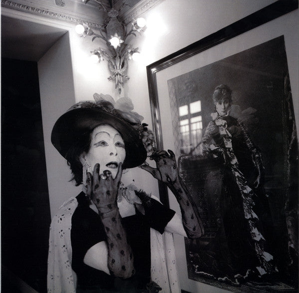 A person with a hat and black veil in heavy white makeup, with a portrait in the background.