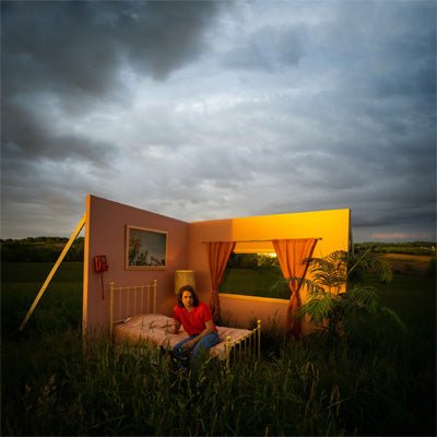 A person sitting in a pink vinyl LP cover depicts a 2 walled open room in a field and a cloudy sky in the background.